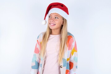 Amazed little kid girl with Christmas hat wearing yarn jacket over white background bitting lip and looking tricky to empty space.