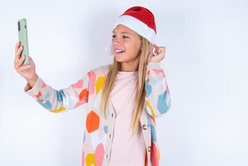 little kid girl with Christmas hat wearing yarn jacket over white background smiling and taking a...