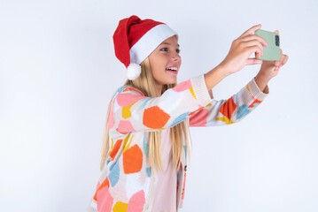 little kid girl with Christmas hat wearing yarn jacket over white background taking a selfie to...