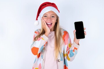 little kid girl with Christmas hat wearing yarn jacket over white background hold hand modern technology use touch face palm astonished impressed scream wow omg unbelievable unexpected