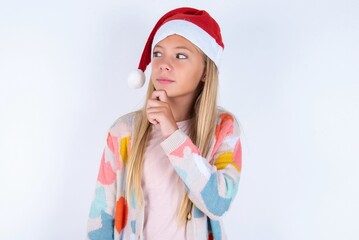 Thoughtful little kid girl with Christmas hat wearing yarn jacket over white background holds chin and looks away pensively makes up great plan