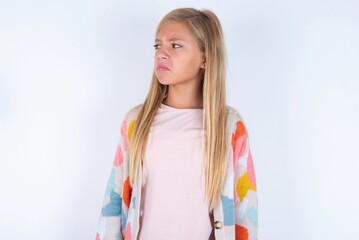 Dissatisfied little kid girl wearing colorful yarn jacket over white background purses lips and has unhappy expression looks away stands offended. Depressed frustrated model.