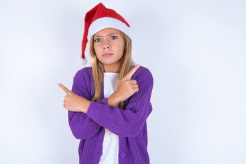 Serious little kid girl with Christmas hat wearing yarn jacket over white background crosses hands and points at different sides hesitates between two items. Hard decision concept