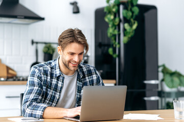 Satisfied caucasian positive young man, stylishly dressed, freelancer, IT specialist, crypto trader, sits in the kitchen at home, working remotely online on a new project, typing on a keyboard, smiles