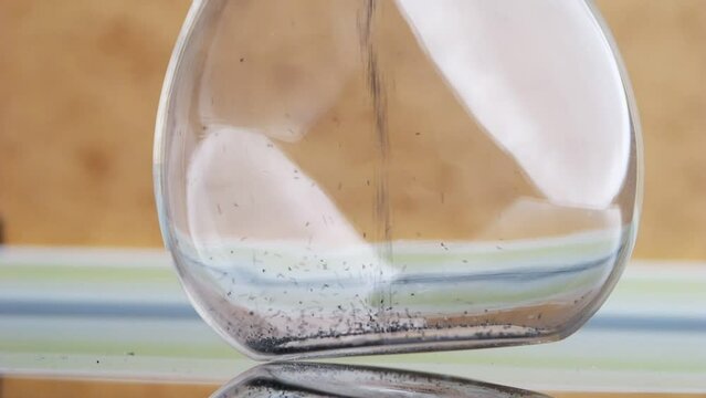 Grains of black sand and quartz are falling in a glass hourglass.