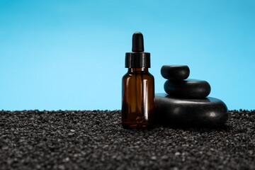 Cosmetic dropper bottle and spa stones on black sand against blue background