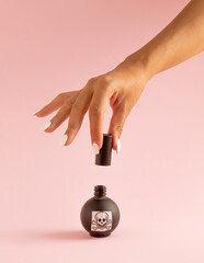 Woman's hand with red nails and poison in black bottle against pastel pink background. Halloween surreal spooky idea.