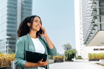 Busy successful curly brazilian or latino woman, formally dressed, realtor, broker, stands outdoors against the background of business center, talking by smart phone with client, looking ahead, smile