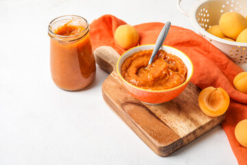 Wooden board with bowl of tasty apricot jam on light background