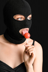 Young woman in balaclava licking lollipop on black background