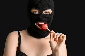 Young woman in balaclava biting lollipop on black background