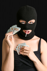Young woman in balaclava with lollipop holding money on black background
