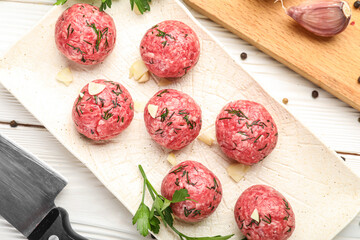 Plate with raw meat balls, herbs, garlic and peppercorns on white wooden background, closeup