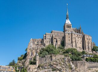 Fototapeta na wymiar Mont St. Michel, Normandy, France - July 8, 2022: Entire gray stone church building with tower, spire, and St. Michael statue on top, stands on its rock surrounded by ramparts under blue sky.
