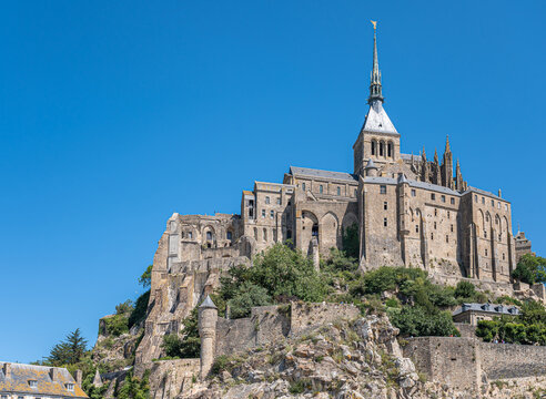 Mont St. Michel, Normandy, France - July 8, 2022: Entire gray stone church building with tower, spire, and St. Michael statue on top, stands on its rock surrounded by ramparts under blue sky.