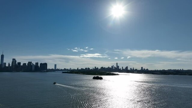 An aerial view of NY harbor on a sunny day. The sun glistens on the water as boats sail by. The camera pan left to show Governors Island, Brooklyn, Queens, lower Manhattan and NJ in the background.