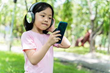 Close up of asian child girl with smartphone and listens to music with modern headphones wireless in park outdoors.