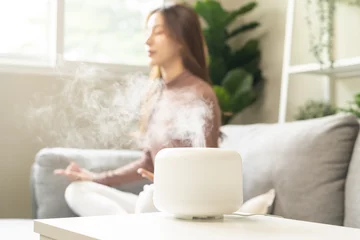 Foto op Plexiglas Air humidifier, calm blurred woman, girl sitting on couch lotus pose put hands practice meditation do yoga exercise at home. Aromatherapy steam scent from essential oil diffuser in living room at home © KMPZZZ