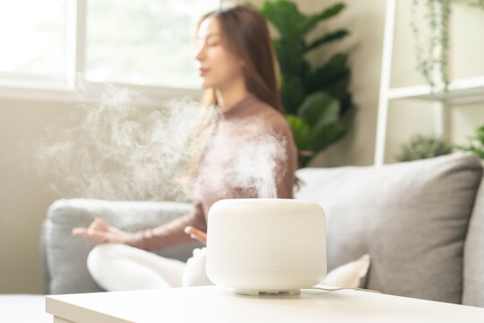 Air humidifier, calm blurred woman, girl sitting on couch lotus pose put hands practice meditation do yoga exercise at home. Aromatherapy steam scent from essential oil diffuser in living room at home