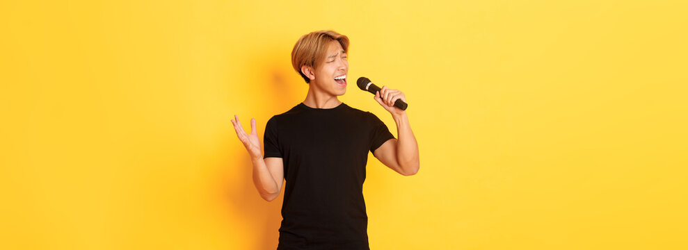 Carefree handsome asian guy, korean singer singing into microphone with passion, standing yellow background