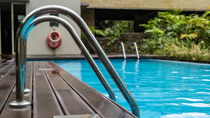 Metal railings are lowered into a swimming pool with blue water. Drops are visible on the handrails. Close-up. In the distance, life buoys hang on the wall. Soft background- tropical vegetation