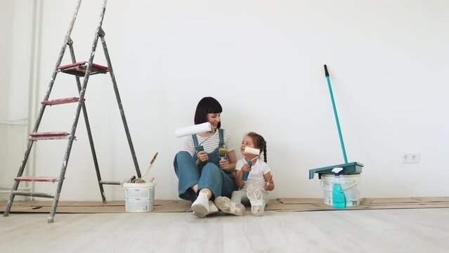 Young mother and her little blonde cute girl with wall paint rollers and brushes sitting on the floor during home renovation on white wall background.