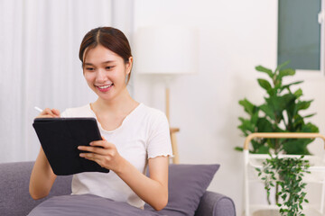Lifestyle in living room concept, Young Asian woman writing data on tablet while sitting on couch