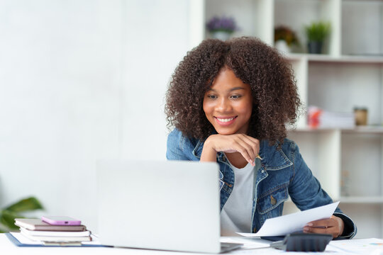 Smiling beautiful business woman happily and comfortably working on her laptop computer and taking notes and working on paperwork at the office..