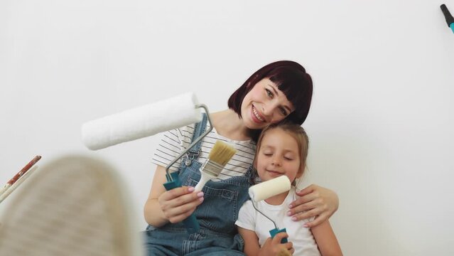 Young mother hugging her little blonde cute girl with wall paint rollers and brushes sitting on the floor during home renovation on white wall background.