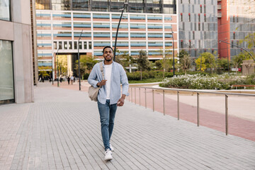 Fototapeta na wymiar Full length handsome young african guy walks through european city during day. Brunette man wears shirt, jeans and backpack. Happy day concept