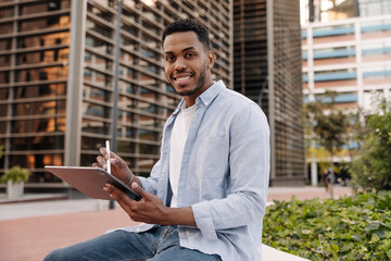 Positive young african guy smiling at camera, holding tablet, stylus sitting on street. Brunette...