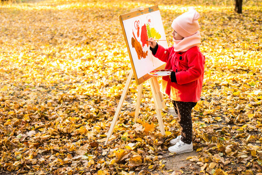 A little girl in a red coat paints a watercolor on the easel in the park against the background of the autumn landscape