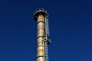 old silver color rusty metal industrial chimney stack. industry and manufacturing concept. grunge detail. exterior access ladder. safety cage. blue sky. Russian gas shortage in Europe concept.
