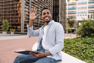 Happy young african man waving to side sitting outdoors with tablet. Brunette guy wears casual...