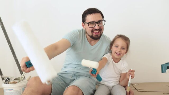 Young father hugging his little blonde cute girl with wall paint rollers and brushes sitting on the floor during home renovation on white wall background.