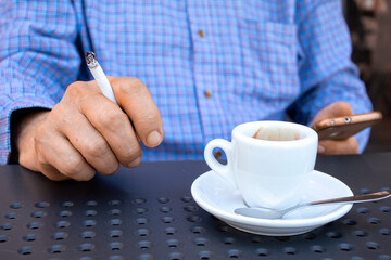 An unrecognisable man is holding a lighted cigarette with one hand while with the other one he is texting. There is a coffee cup next to him. He is sitting at a cafeteria table