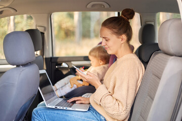 Side view portrait of puzzled helpless woman working on laptop while sitting with her baby daughter in safety chair on backseat of the car, using cell phone with confused expression.