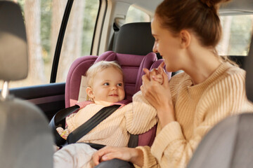 Mother with her funny little daughter in safety chair in the car, to ride in a family auto, playing around, mommy kissing or biting baby's fingers, or kid pointing to her mom mouth.