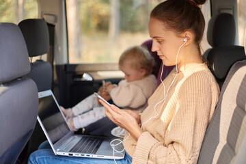 Profile portrait of serious woman with earphones and mobile phone working on laptop while sitting with her toddler kid in safety chair on backseat of the car.