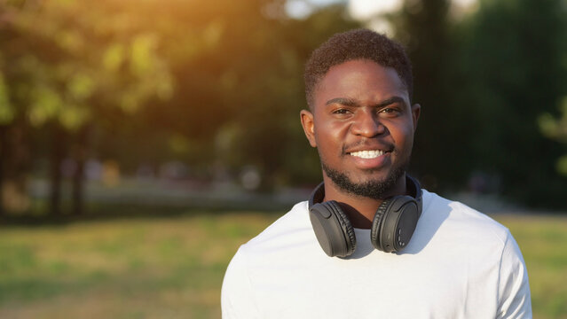 Smiling African American man wearing black headphones walks in green park. Black adult enjoys walking exploring great destination at sunlight