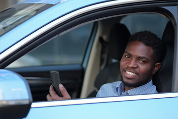 African American driver sitting at car steering wheel talks with friends via videocall. Bearded man spends time in traffic jams communicating with friends