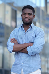 Stylish African American businessman with arms folded in blue shirt stands and smiles at camera against office building.