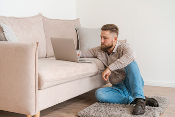 Young attractive, smiling bearded guy browsing his laptop while sitting at home on a cozy beige sofa at home, in casual clothes on a day off