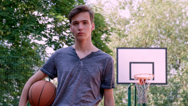 Teenager holds a basketball in his hand is on the sport ground and looking to the camera. Handsome Young man on a basketball ground with a basketball in his hands. Sport and activity concept.