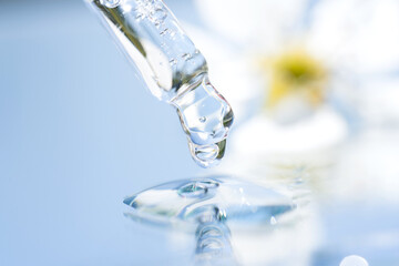 Dripping drop of transparent liquid with glass pipette against light blue background with flowers. Texture of face serum, herbal essence or natural cosmetic oil. Selective focus