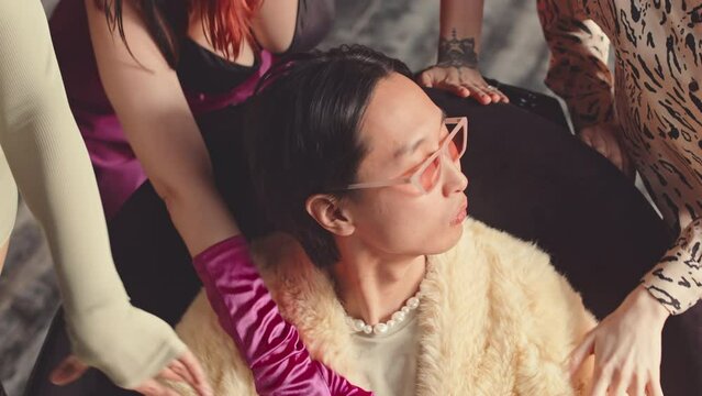 Slowmo of unrecognizable girls dancing seductively around pompous Asian guy in retro pink eyeglasses and fur coat sitting on chair examining them