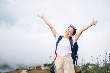 Young smiling Asian woman with backpack enjoying travel. Beautiful tourist female raised hands with background of fog and village on mountain. Freedom and happy life concept.
