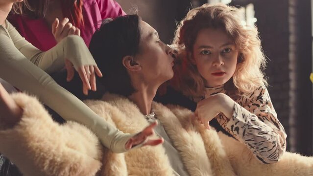 Slowmo of confident Asian guy in camel fur coat looking at camera pompously while sitting in chair among three beautiful fancy girls dancing vogue to him