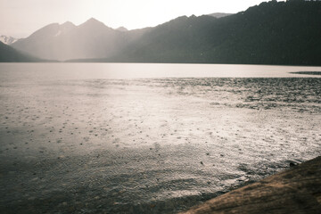 Rain on the surface of a mountain lake. Mountain range silhouette in the background. Sun during the rain.