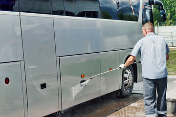 Car wash worker manually washes large bus with stream of water in open parking lot on summer day. Service for washing large equipment. Authentic scene workflow.